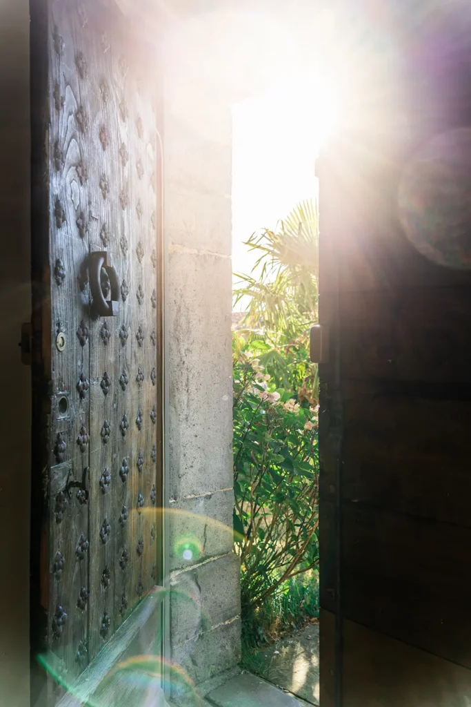 Porte ancienne ouvrant sur un jardin avec palmier et soleil, Maison Belzunce par Studio ADIO, Pays Basque.