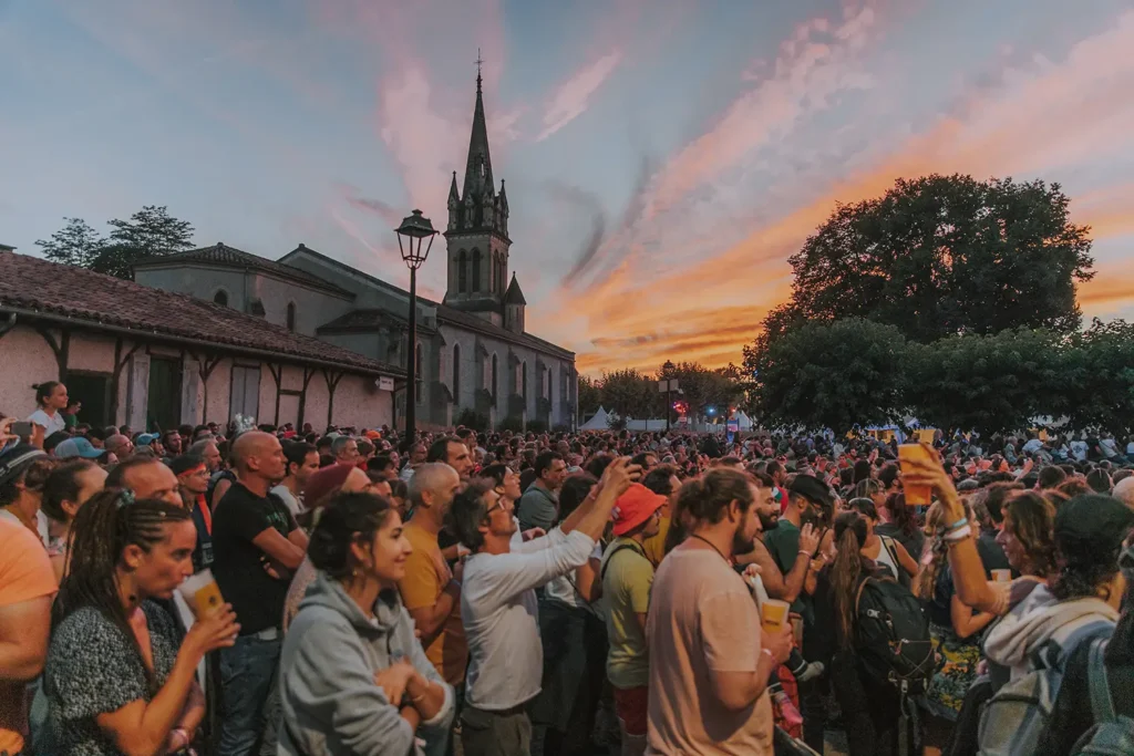 Photo d'ambiance : foule devant l'église de Luxey sous un coucher de soleil coloré au festival Musicalarue par l'agence ADIO