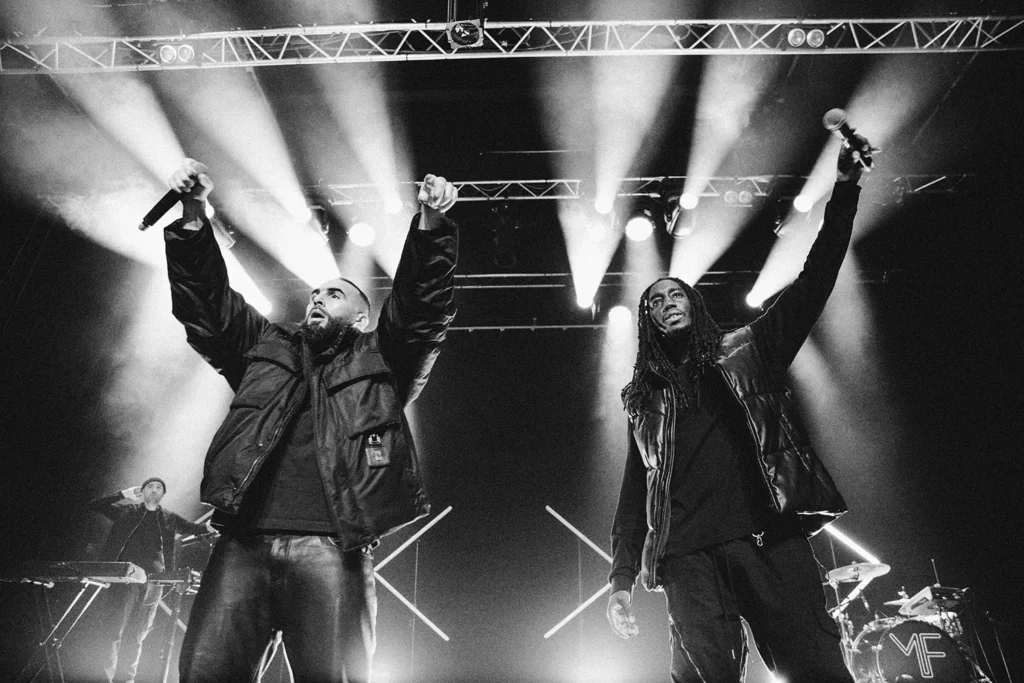Photographie de scène : Médine et son accompagnateur les bras levés face au public en noir et blanc à l'Atabal par l'agence ADIO